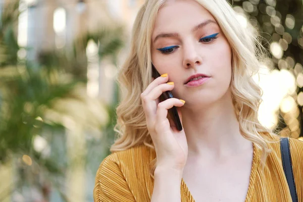 Portrait of serious blond girl with makeup thoughtfully talking on cellphone outdoor — Stock Photo, Image