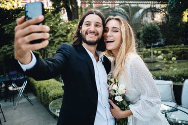 Genç çekici damat ve gelin sevinçle restoranın güzel bahçesinde birlikte cep telefonu selfie alarak 