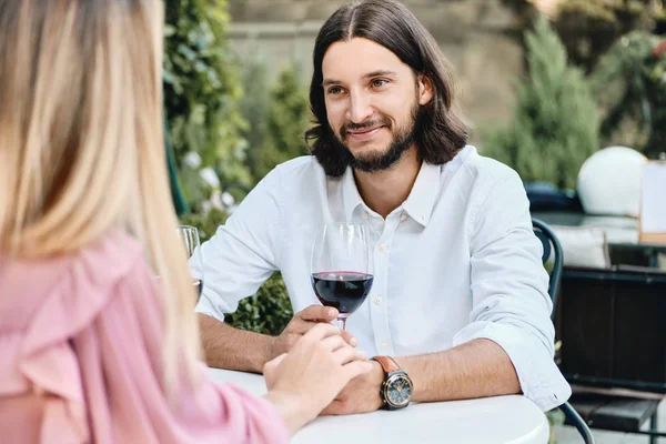 Joven guapo morena barbudo hombre en camisa con copa de vino felizmente mirando novia en cita romántica en la cafetería al aire libre — Foto de Stock