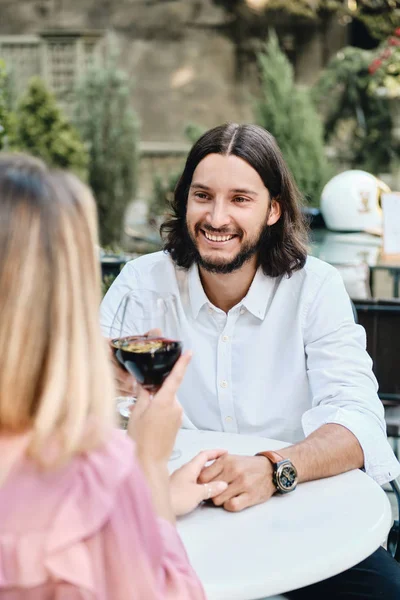 Joven atractiva morena barbuda hombre en camisa con copa de vino mirando alegremente a la novia en una cita romántica en la cafetería al aire libre — Foto de Stock