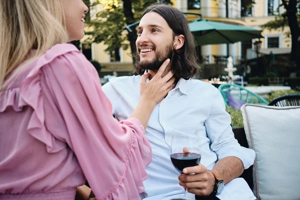 Joven guapo morena barbudo hombre en camisa con copa de vino felizmente mirando novia en romántica fecha en restaurante al aire libre — Foto de Stock