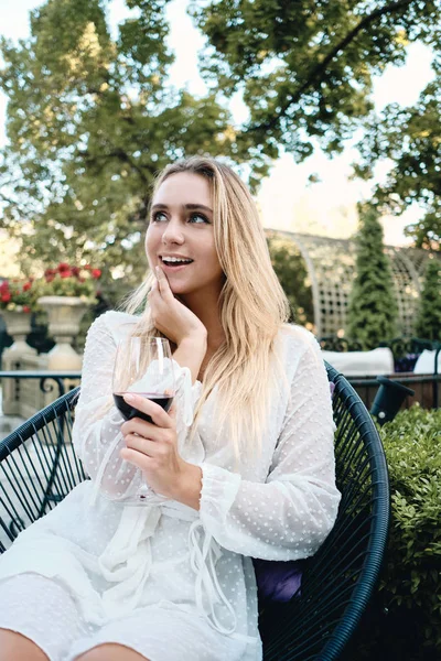 Young gorgeous blond woman in white dress dreamily looking away with glass of wine in garden of restaurant — Stock Photo, Image