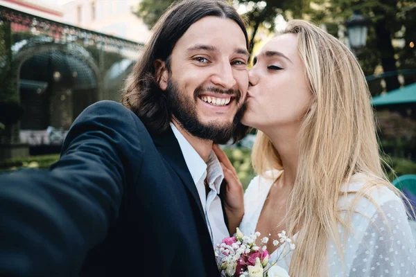 Νέος ελκυστικός χαμογελαστός γαμπρός με χαρά λήψη selfie, ενώ όμορφη νύφη φιλιά του στο μάγουλο εξωτερική — Φωτογραφία Αρχείου
