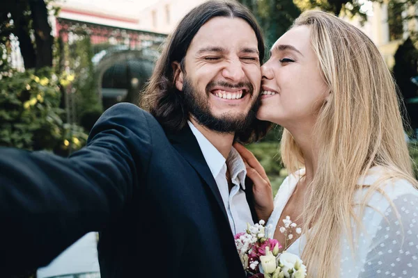 Νέος ελκυστικός συναισθηματικός γαμπρός με χαρά λήψη selfie, ενώ όμορφη νύφη φιλιά του στο μάγουλο στον κήπο του εστιατορίου — Φωτογραφία Αρχείου