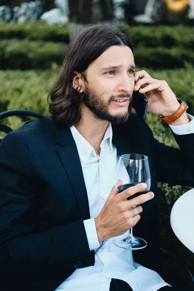 Giovane attraente bruna barbuto uomo d'affari con un bicchiere di vino pensieroso guardando lontano parlando sul cellulare in ristorante all'aperto — Foto Stock
