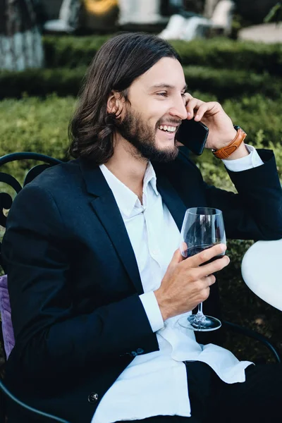 Giovane bruna sorridente uomo d'affari barbuto con un bicchiere di vino che parla felicemente sul cellulare nel ristorante all'aperto — Foto Stock