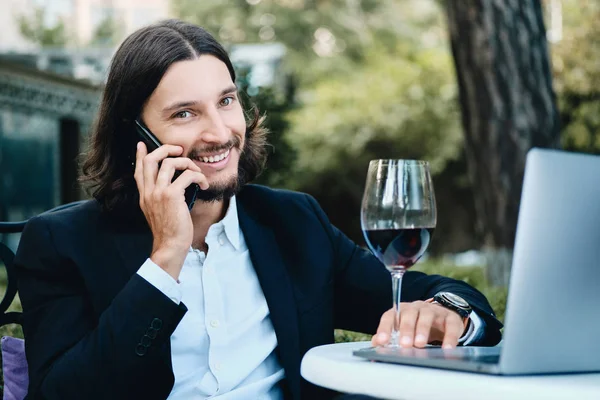 Junge hübsche brünette bärtige Geschäftsmann mit einem Glas Wein glücklich in die Kamera im Gespräch auf dem Handy im Restaurant im Freien — Stockfoto