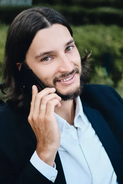 Young handsome confident bearded businessman joyfully looking in camera talking on cellphone in restaurant outdoor — Stock Photo, Image