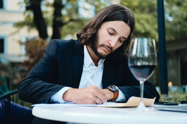 Giovane bell'uomo d'affari serio barbuto con un bicchiere di vino che lavora premurosamente nel ristorante all'aperto — Foto Stock