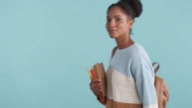 Sırt çantalı ve not defterli güzel Afrikalı Amerikalı kız öğrenci mavi arka planda kameraya sevinçle bakıyor.