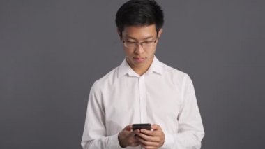 Gözlüklü yakışıklı Asyalı genç adam gri arka planda cep telefonu kullanıyor. 