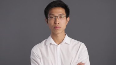 Gözlüklü Asyalı genç adam gözlerini deviriyor ve kabahatli bir şekilde gri arka plana bakıyor. 