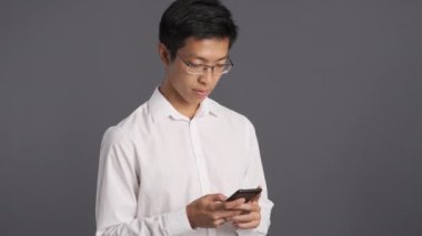 Gözlüklü genç ve çekici Asyalı adamın gri arka planda cep telefonunu mutlu bir şekilde kullanması. 