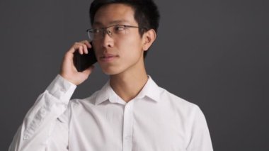 Gözlüklü genç ve çekici Asyalı adam cep telefonuyla sevinçle konuşuyor ve gri arka planda zafer işareti yapıyor. 