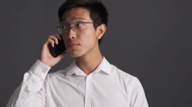 Genç üzgün gözlüklü Asyalı adam gri arka planda cep telefonuyla konuşuyor. 