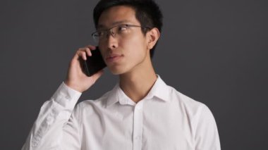 Gözlüklü Asyalı genç adam gri arka planda can sıkıcı cep telefonuyla konuşuyor. 