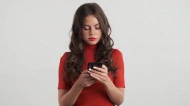 Kırmızı elbiseli, esmer, güzel bir kız. Gri arka planda cep telefonunu incelikle kullanıyor.