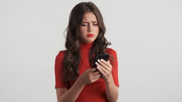 在灰蒙蒙的背景下使用手机 令人不快的黑发女孩 穿着红色上衣 很失望 — 图库视频影像