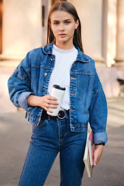 Kot ceketli, şık, sıradan bir kız öğrenci. Açık havada kameraya bakıyor.