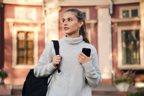 Attraente studentessa casual in piedi con zaino e cellulare all'aperto — Foto Stock