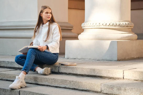 Menina estudante casual atraente estudando cuidadosamente com livro em escadas da universidade ao ar livre — Fotografia de Stock