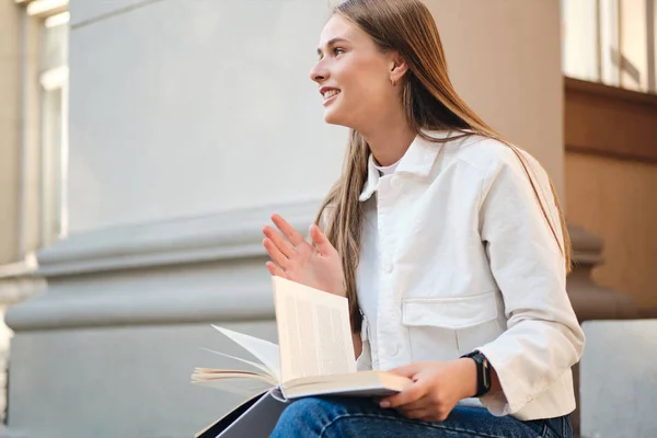 Vista lateral da menina estudante casual alegre se preparando para estudar com livro durante a pausa na universidade ao ar livre — Fotografia de Stock