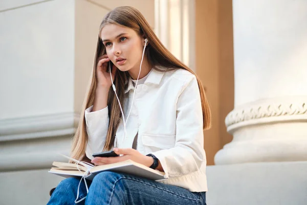 Piękny casual uczennica w słuchawki starannie słuchanie muzyki na telefon komórkowy podczas przerwy w uczelni odkryty — Zdjęcie stockowe