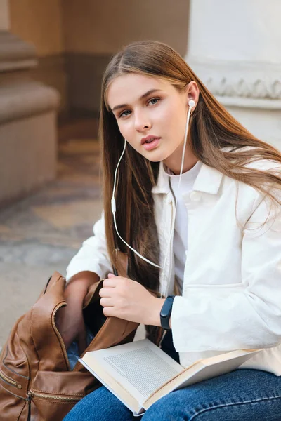 Menina estudante casual bonita em fones de ouvido com livro olhando atenciosamente na câmera durante a pausa na universidade ao ar livre — Fotografia de Stock