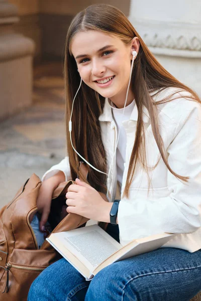 Melko hymyilevä rento opiskelija tyttö kuulokkeet kirjan iloisesti etsivät kameran tauon aikana yliopiston ulkona — kuvapankkivalokuva