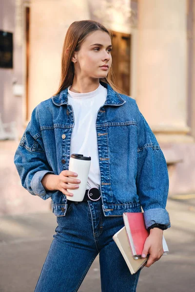 Attraktiv snygg casual student flicka i denim jacka med kaffe och läroböcker eftertänksamt tittar bort utomhus — Stockfoto