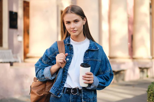 Krásná stylová ležérní studentka v džínové bundě s batohem zasněně dívá do kamery venku — Stock fotografie