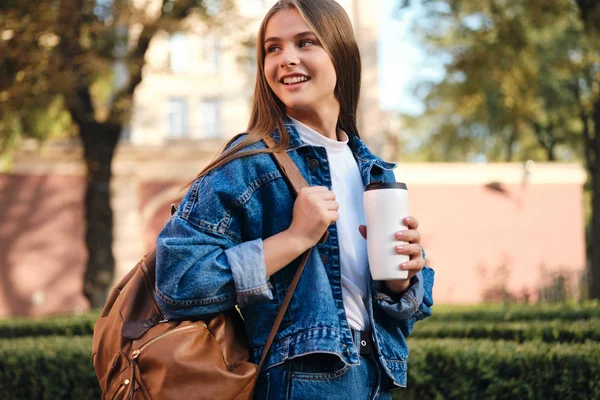 Elég vidám alkalmi diák lány farmerdzsekiben, hátizsákkal és kávéval boldogan néz félre a főiskolai campus outdoo-ban. — Stock Fotó