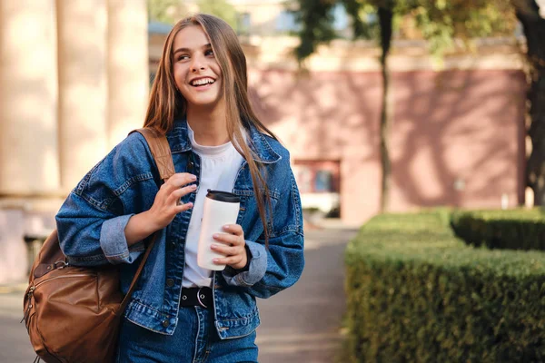 Glada casual student flicka i denim jacka med ryggsäck glatt tittar bort i universitetet utomhus — Stockfoto