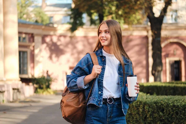 Całkiem uśmiechnięta studentka w dżinsowej kurtce z plecakiem i kawą na przerwie w studiach w college 'u outdoo — Zdjęcie stockowe