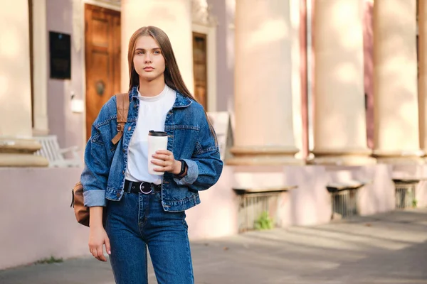 Gyönyörű alkalmi diák lány farmerdzsekiben, hátizsákkal és kávéval figyelmesen néz félre az egyetemen szabadban — Stock Fotó