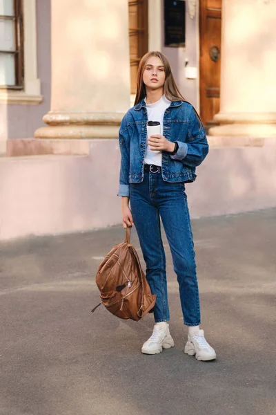 Bella ragazza studentessa casual elegante in giacca di jeans con zaino guardando fiduciosamente in macchina fotografica in università all'aperto — Foto Stock