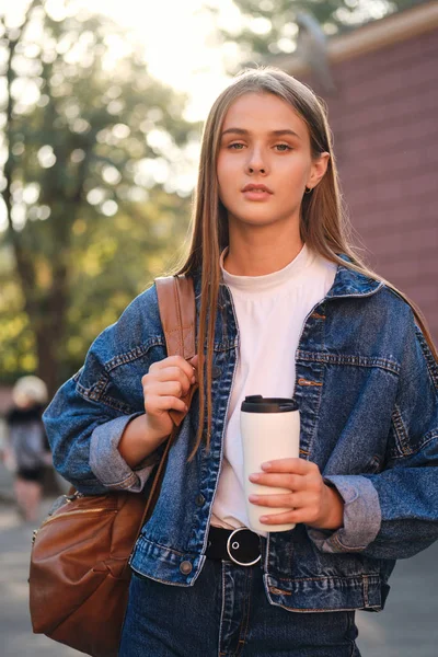 Jovem menina estudante casual lindo em jaqueta de ganga com mochila e copo para ir pensativo olhando na câmera ao ar livre — Fotografia de Stock