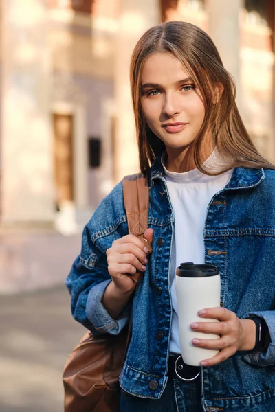 Ung ganska avslappnad student flicka i denim jacka med kopp för att gå drömskt tittar i kameran i universitetet utomhus — Stockfoto