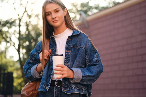 Ung vacker casual student flicka i denim jacka med ryggsäck och kopp för att gå drömskt tittar i kameran utomhus — Stockfoto
