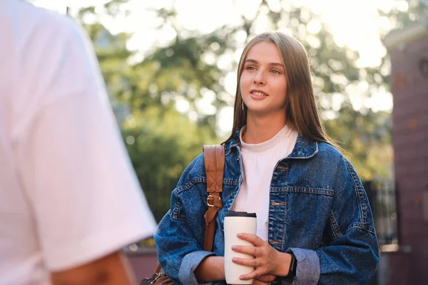 Pěkně ležérní studentka v džínové bundě s šálkem jít zasněně mluvit s přítelem na studijní přestávce na vysoké škole venkovní — Stock fotografie