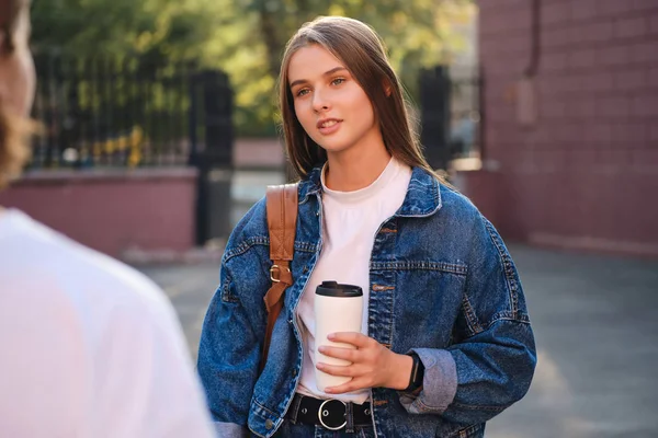 Giovane attraente studentessa casual in giacca di jeans con coppa per andare a parlare con un amico in pausa studio in università all'aperto — Foto Stock