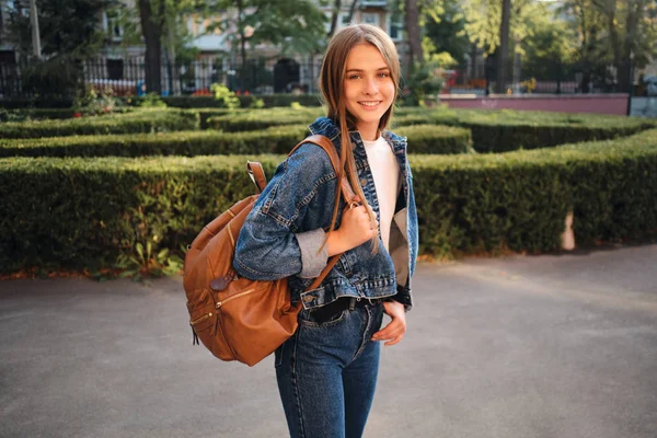 Krásná usměvavá ležérní dívka v džínové bundě se stylovým batohem radostně dívá do kamery venku — Stock fotografie