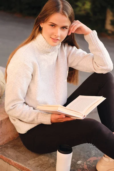 Молодая красивая студентка в уютном свитере счастливо смотрит в камеру, учится с учебником под открытым небом — стоковое фото