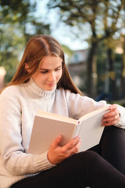 Молодая красивая студентка в уютном свитере мечтательно читает книгу на открытом воздухе — стоковое фото