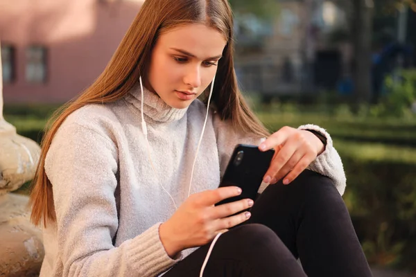 Gadis pelajar muda yang menarik dengan sweater yang nyaman dengan menggunakan ponsel selama istirahat belajar di luar ruangan — Stok Foto