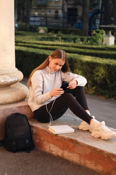 Fiatal gyönyörű, elegáns diáklány kényelmes pulóverben álmodozva használja a mobilt a tanulmányi szünet alatt szabadtéri — Stock Fotó