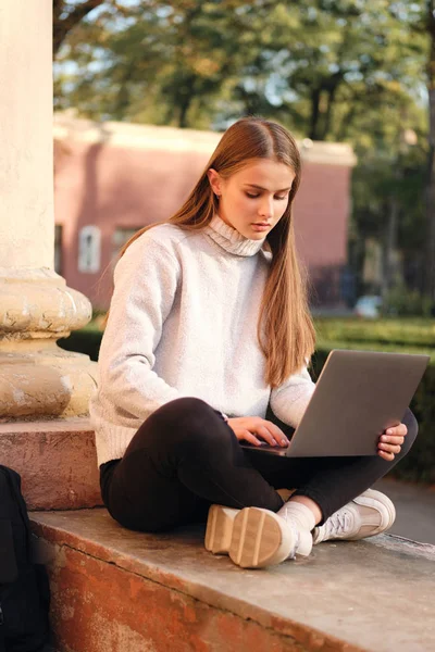 Muda cantik berambut coklat gadis mahasiswa dalam sweater nyaman belajar dengan laptop outdoor — Stok Foto