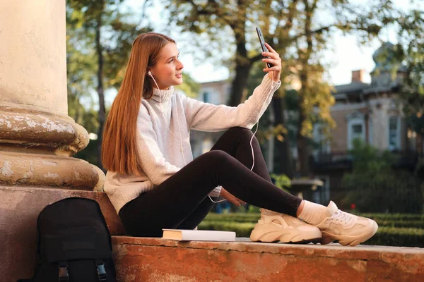 Gadis muda yang menarik dengan sweater yang nyaman bermimpi mengambil foto selfie di ponsel selama istirahat belajar di luar ruangan — Stok Foto