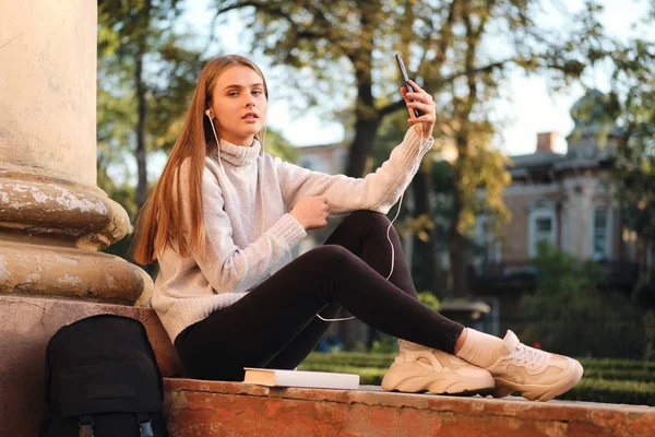 Vonzó diáklány kényelmes pulóverben figyelmesen nézi a kamerát, ahogy szelfit készít a mobilján a tanulmányi szünetben a szabadban — Stock Fotó