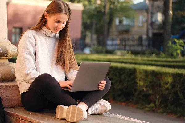 Joven bastante sonriente chica estudiante de cabello castaño en suéter acogedor estudio de ensueño con ordenador portátil al aire libre —  Fotos de Stock